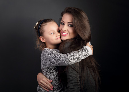 Happy mother hugging her fun daughter on dark grey background in studio with loveの写真素材