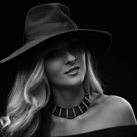 Beautiful expravagant makeup woman posing in fashion blue hat and gold necklace on dark black background. Black and white portraitの写真素材