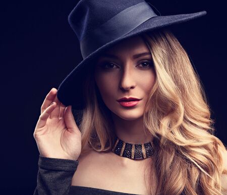 Sexy makeup blond long hair style woman posing in fashion hat and gold necklace on dark background....