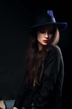 Expressive female makeup model posing in black shirt and elegant hat with red lipstick on dark background and with thinking look looking down. Dramatic lightの写真素材