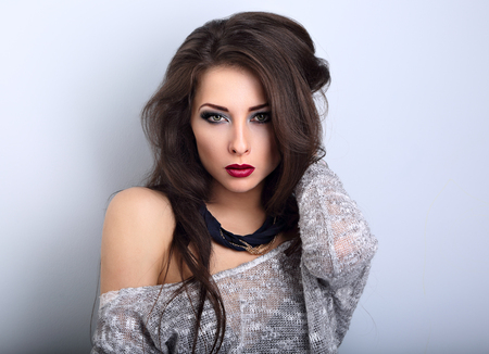 Beautiful young expressive make-up model posing in grey blouse with modern necklace on the neck on blue background with empty copy space. Long hairstyle and red lipstick.の写真素材