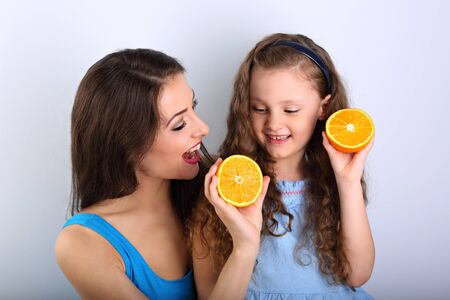 Enjoying fun young mother showing her cute long hair daughter the slices of fresh bright orange with happy toothy smiling. Healthy and diet lifestyle. の写真素材