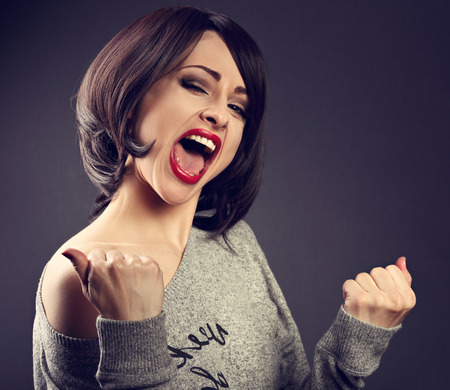 Excited winner makeup short hair style woman with opened mouth. Happy young satisfacted female showing the gesture yes by fists on grey dark background. Closeup toned portraitの写真素材