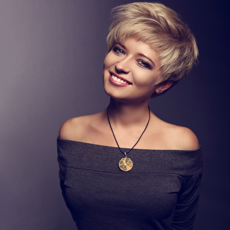 Happy toothy smiling woman with short blond bob hairstyle in grey blouse and orange fashion necklace on gray background. Closeup toned portraitの写真素材