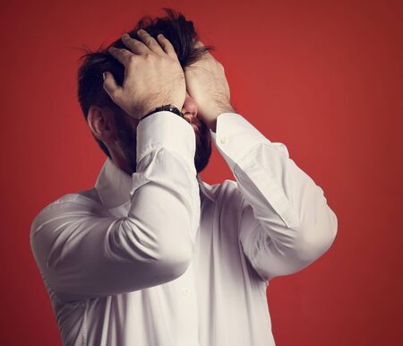 Unhappy bearded business man covering face his hands in white shirt on red background. Closeup toned...