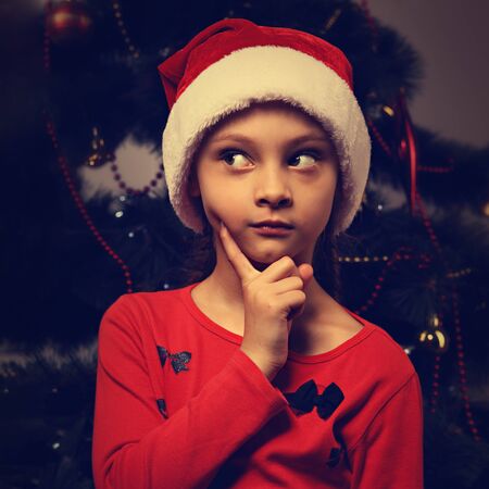 Cute kid girl with in Christmas santa claus hat thinking about something enigmatic and holding the finger under the face on green fur tree background. Closeup toned bright art portraitの写真素材