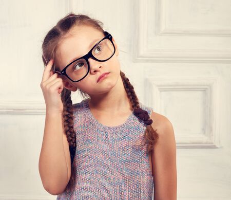 Happy thinking kid girl in fashion glasses and blouse with serious emotional face scratching the head. Toned closeup portraitの写真素材