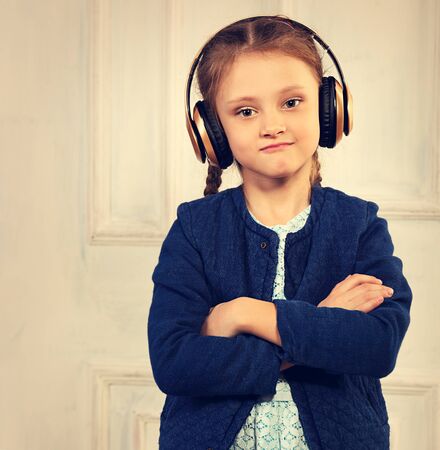 Serious kid girl in blue jacket listening the music in wireless headphone with folded arms on studio background. Toned vintage portraitの写真素材