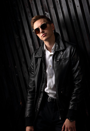 Sexy fashion relaxing male model posing in leather jacket and trendy eyeglasses on black studio background. Closeup portraitの写真素材