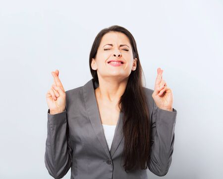 Beautiful smiling business woman in office clothing making a wish and crossing the fingers with squinting eyes and looking up on blue background. Closeup studio portraitの写真素材