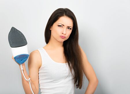 Fun attractive unhappy aggressive woman holding the home iron and wanting to hit on grey background. Closeup portraitの写真素材