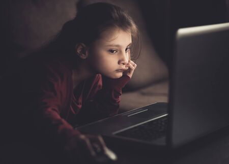 Unhappy small kid pupil girl learning and looking on computer monitor at home. Dark shadow closeup vintage toned portrait.の写真素材