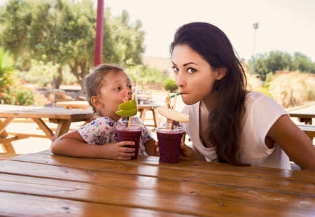 Happy kid girl and grimacing emotional mother drinking berries smoothie juice together in street summer cafe. Closeup portraitの写真素材