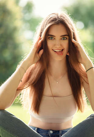 Beautiful smiling teen woman looking happy and making funny mug and showing the tongue sitting on the green glass outdoors summer green trees background. Closeup portrait in sunny dayの写真素材