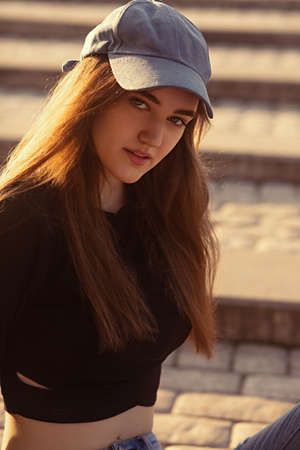 Beautiful candid smiling woman in blue cap looking sexy outdoors summer orange sunset background. Closeup portrait in bright sunny day autumn day. Toned brightの写真素材