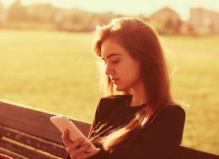 Happy smiling teen girl sitting on autumn background in sunny day and using the mobile phone. Closeup portriatの写真素材