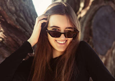 Beautiful toothy laughing woman looking happy in fashion sunglasses outdoors summer green trees background. Closeup portrait in bright sunny dayの写真素材
