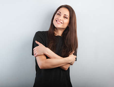 Beautiful casual cute toothy smiling woman hugging your self with folded strong arms in black casual t-shirt on blue background. Closeup portrait with empty copy spaceの写真素材
