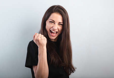Happy excited beautiful woman showing success triumph sign by fist and loud shouting with open mouth on blue background with empty copy space. Closeup portraitの写真素材