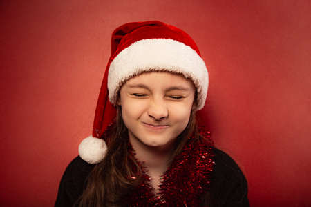 Fun child girl screw up one's eyes guessing the wish in santa claus hat on red background. Happy Christmas closeup portraitの写真素材