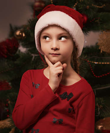 Cute kid girl with in Christmas santa claus hat thinking about new year gift and holding the finger under the face on green fur tree background. Closeup toned portraitの写真素材