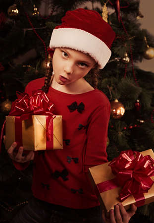 Cute excited dancing unhappy kid girl in Christmas santa claus hat holding the red box with gift with expression surprising face on green fur tree background. Closeup portraitの写真素材