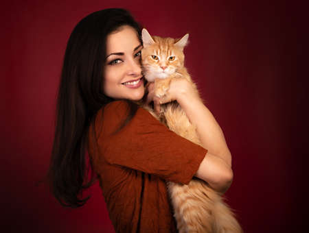 Beautiful happy smiling young woman holding and hugging with love her red maine coon kitten. Closeup portrait on red holiday background with empty copy spaceの写真素材