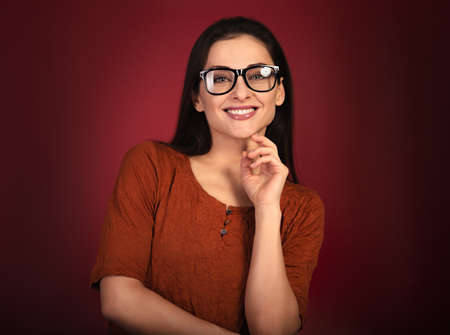 Beautiful thinking toothy smiling happy woman looking in eye glasses with hand under the face in casual orange t-shirt on red background with empty copy space. Closeup portrait.の写真素材