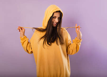 Relaxing fun happy smiling woman posing in fashion yellow hoodie with hood on the head on purple bright background with empty copy space. happy portraitの写真素材
