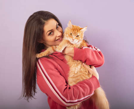 Beautiful happy smiling young woman holding on the hands and hugging with love her red maine coon kitten. Close up portrait on purple background with empty copy spaceの写真素材