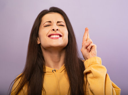 Squint smiling young desiring woman in yellow hoodie crossing fingers with closed eyes on purple bright background. close-up portraitの写真素材