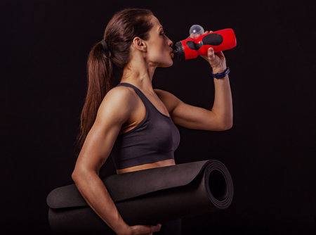 Happy pretty fit healthy fitness woman holding yoga mat and drinking from sport flask on black background with empty copy space. Healthy lifestyle portraitの写真素材