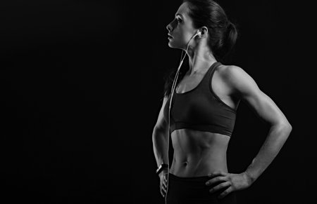 Profile shot of serious sporty female with ponytail, listening music in headphones in sportsbra, standing isolated on black background with empty copy space. Lifestyle sport portrait. Black and whiteの写真素材