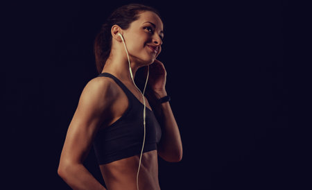Profile shot of happy smile female athlete with ponytail, listening music in headphones, workout in gym in sportsbra, standing on black background with empty copy space. Lifestyle portrait training in earphones. Toned colorの写真素材