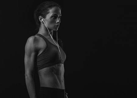 Profile shot of serious female athlete with ponytail, listening music in headphones in sportsbra, standing isolated on black background with empty copy space. Lifestyle portrait in earphones. Black and whiteの写真素材