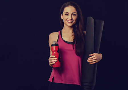 Happy pretty fit healthy fitness woman in pink sporty top clothing holding black mat for yoga and red sporty flask on black background with empty copy space. Healthy lifestyle portrait. Toned colorの写真素材