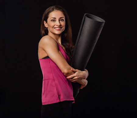 Happy pretty fit healthy fitness woman in pink sporty top clothing holding and hugging black mat for yoga on black background with empty copy space. Healthy lifestyle positive portrait.の写真素材