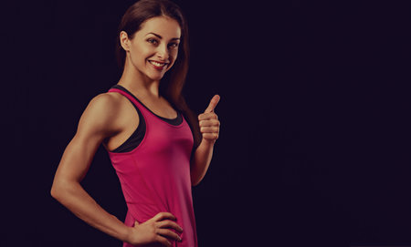 Happy smiling sportswoman in pink sportsbra and shirt, fitness trainer showing thumb up sigh by hand standing isolated on black background with empty copy space. Closeup banner portrait. Toned colorの写真素材