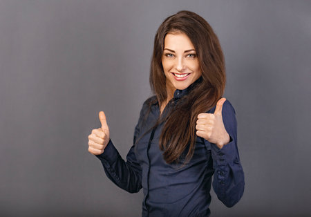 Beautiful excited energetic smiling business woman showing fingers thumb up by two hands, the ok sign in blue shirt on gray background. close-up portraitの写真素材