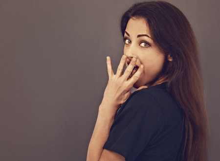 Surprised beautiful woman looking with big eyes, opened mouth and covering mouth the hand in black shirt on gray background. Closeup toned color portraitの写真素材