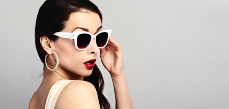 Fashion studio closeup portrait of gorgeous elegant surprising woman in white sunglasses and fashion earrings posing on wall background with empty copy space. Banner portraitの写真素材