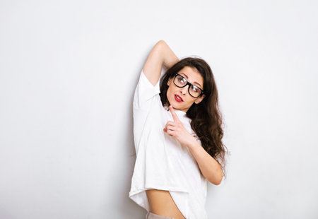 Beautiful fun makeup woman in eyeglasses in white casual t-shirt looking and showing up and demonstrating idea sign by finger above on empty copy space white studio background.の写真素材