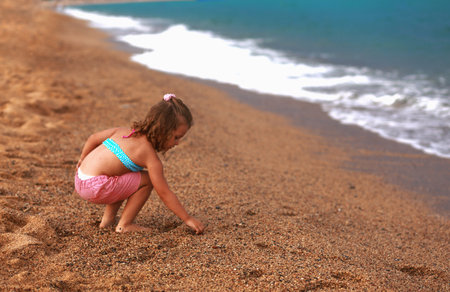 Beautiful fun happy kid girl sitting in summer clothing on the yellow sand and playong with stones of the beach near blue sea and looking on water background. Happy vacationの写真素材