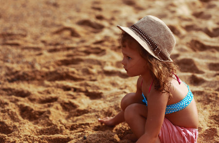 Beautiful happy kid girl thinking and sitting in summer clothing, straw hat on the yellow sand of the beach and looking on water background. Happy vacation closeup portraitの写真素材