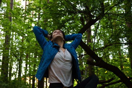Beauty woman making the arms fly sign, looking up in the pine forest. Female sitting in sport clothes in a park with arms behind head. Girl breath deep in green woodの写真素材