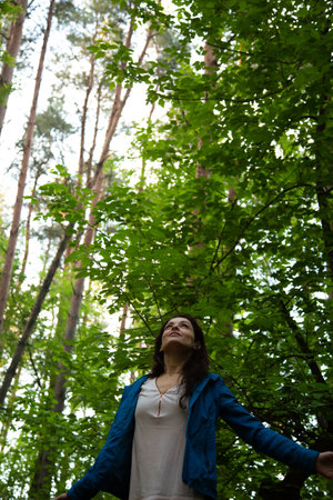 Beauty woman making the arms fly sign, looking up in the pine forest. Female gesture free sign in a park with arms raised back. Girl breath in green wood with trees aboveの写真素材
