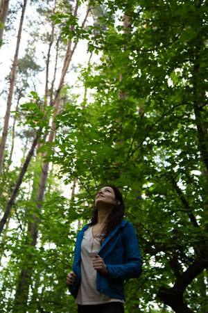 Beauty woman looking up in the pine forest. Female standing in blue style coat in a park and looking...