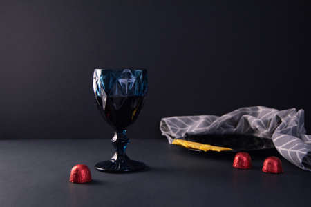 Blue glass of red wine on a black background. Pieces of cheese on a black plate, a gray kitchen napkin and candies in a red packageの写真素材