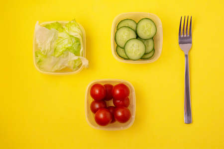 Healthy food. Delivery of products. Lettuce, cucumber slices, tomatoes: vegetables in separate containers and a fork on a yellow backgroundの写真素材