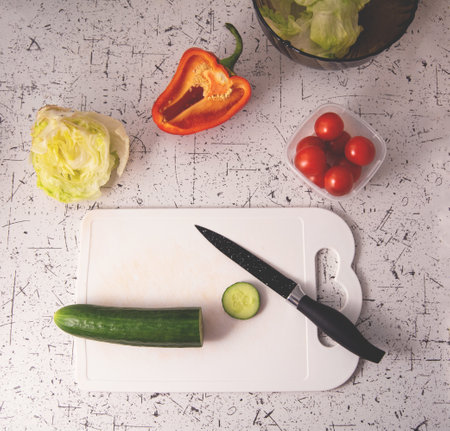 Ingredients for preparing salad on the kitchen table. Salad bowl with lettuce, red tomatoes, green cucumbers and lettuce. Knife on a cutting boardの写真素材